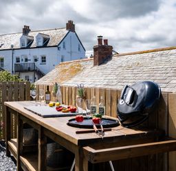 An outdoor wooden table with glass bottles, wine glasses, vegetables, and a black grill lid on a fenced patio at Mino Cottage in Port Isaac