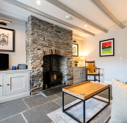 A living room with a stone fireplace a wooden coffee table a TV a chair with a cushion and framed artwork on the walls at Mino Cottage in Port Isaac