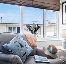 A gray armchair with a blue nautical pillow a hat a scarf and a book by a window with flowers and a table lamp at Mino Cottage in Port Isaac
