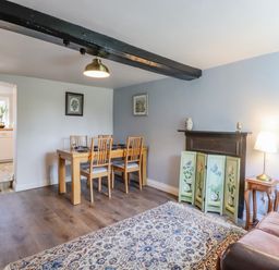 A dining area with a wooden table and four chairs next to a living room with a patterned rug a brown sofa and a small wooden side table with a lamp at Bluebird Cottage in Newmarket
