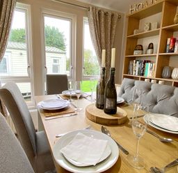 A dining area with a wooden table set with plates glasses and cutlery with chairs and a cushioned bench near windows and a bookshelf with books and decorative items at Meadow Lodge in Cayton