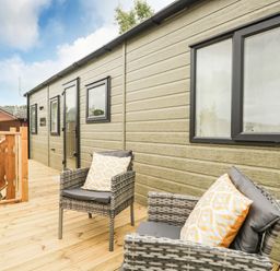 An outdoor wooden deck with two wicker chairs with cushions next to a green building with black windows at Ashton Lodge - Sherwood 4 in South Lakeland Leisure Village