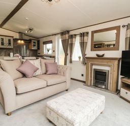 A living room with a beige sofa purple cushions an ottoman a fireplace a flat screen tv and kitchen cabinets at Ashton Lodge - Sherwood 4 South Lakeland Leisure Village