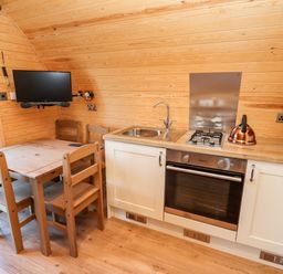A small kitchen with a wooden dining table and chairs a wall mounted TV a sink a gas stove an oven and a copper kettle at Goose Pod Near Clifton Upon Teme