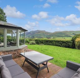 An outdoor patio with wicker sofas and a glass table overlooking green hills at Flaska House in Troutbeck Near Penrith