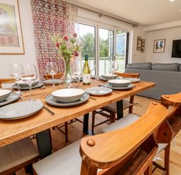 A dining table set with plates glasses and a bottle of wine in a room with wooden chairs a sofa and a wall mounted television at Syrah Seaview
