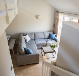A living room with a gray sectional sofa blue and beige cushions a wooden coffee table with flowers and magazines and curtains at 5 Keeper's Cottage Hillfield in Dartmouth