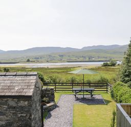 A garden with a picnic table and green umbrella near a stone building and overlooking a grassy field with hills beyond at Minffordd in Trawsfynydd