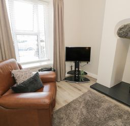 A living area with a brown leather armchair, pillows, a TV on a stand, a window with blinds and curtains, and a fireplace with an electric stove at Minffordd in Trawsfynydd