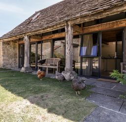 A stone and wood cottage exterior with glass doors and chickens on the grass at Tinkley Cottage in Nympsfield near Nailsworth