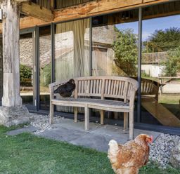 An outdoor wooden bench with a black chicken sitting on it and a brown chicken on the grass in front of glass doors at Tinkley Cottage in Nympsfield near Nailsworth