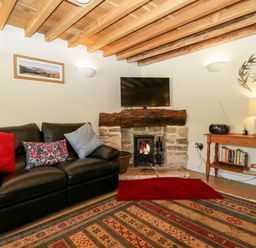 A living room with a black leather sofa cushions a fireplace with a fire a mounted television wooden ceiling beams and a wooden table with books and a lamp at Tinkley Cottage in Nympsfield near Nailsworth