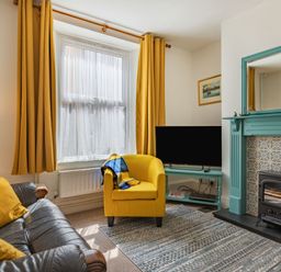 A living room with yellow curtains and armchair a black sofa and a teal fireplace at Sea Spray in Watchet