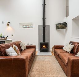 A living room with two brown leather sofas facing a wood burning stove and a television mounted on the wall at Yr Hen Oriel in Llanrug