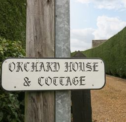 A wooden post with a sign reading Orchard House and Cottage on a gravel path with hedges on either side at Orchard House Cottage in Malmesbury