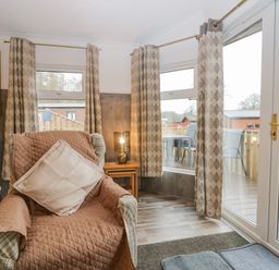 A living room with a chair covered by a quilted cover and cushion a side table with a lamp curtains on windows and glass doors at Wansfell View in Limefitt Park near Troutbeck
