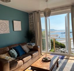 A living room with a brown leather sofa and cushions next to large open doors overlooking a beach and water at Windycroft in Barmouth