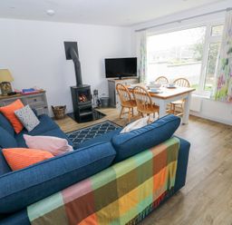 A living room with a blue sofa and colorful cushions a wood burning stove a dining table with chairs and tableware a television and large window with floral curtains at Chalfonts Cottage in Trefin near Penparc