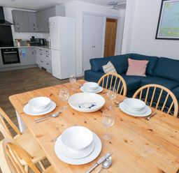 A dining table set with plates bowls and glasses next to a blue sofa and a modern kitchen in the background at Chalfonts Cottage in Trefin near Penparc