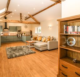An open plan kitchen and living area with green cabinets a gray sofa with pillows a wooden bookshelf and exposed ceiling beams at Y Felin The Granary in Caernarfon