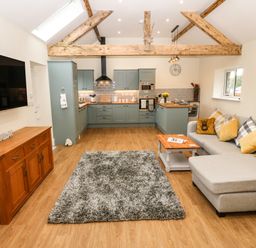 A living room and kitchen area with a gray sofa a wooden coffee table a wooden sideboard a wall-mounted TV and exposed wooden beams at Y Felin The Granary in Caernarfon