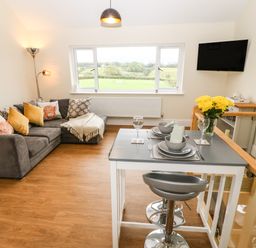A living room with a gray sofa yellow pillows a dining table set for two and a wall-mounted TV at Ty Gwair The Hayloft in Caernarfon