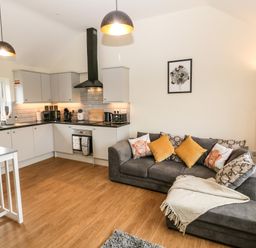 A kitchen with gray cabinets and appliances next to a living area with a gray sofa and yellow and patterned cushions at Ty Gwair The Hayloft in Caernarfon