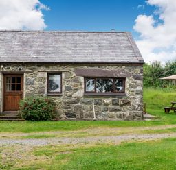 A stone cottage with two windows and a wooden door with bushes in front and a picnic table with an umbrella in the grass at Tryfan Cottage in Rhostryfan near Caernarfon