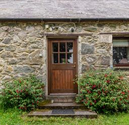 A stone exterior wall with a wooden door and a window surrounded by flowering bushes at Tryfan Cottage in Rhostryfan near Caernarfon