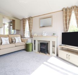 A living room with a beige sofa with cushions a small green ottoman a fireplace with decor a wall mirror and a television on a stand at 25 Sherwood in South Lakeland Leisure Village