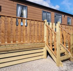 A wooden porch with stairs leading to a door of a brown cabin with windows at 25 Sherwood in South Lakeland Leisure Village