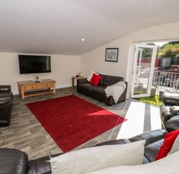 A living room with leather sofas a red rug a wooden TV stand and an open door leading outside at The Apartment at Plas Yr Eithin in Amlwch near Llanfechell
