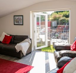 A living room with leather sofas and red cushions by an open glass door leading to an outdoor area at The Apartment at Plas Yr Eithin in Amlwch near Llanfechell