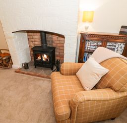 A living room with a wood burning stove a wicker basket with firewood a patterned armchair and a wooden cabinet with a lamp and books at Bumble Cottage in Tysoe