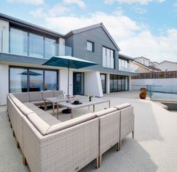 An outdoor patio area with a large cushioned seating arrangement under an umbrella in front of a modern two-story house with glass balcony at Megizzy in Polzeath