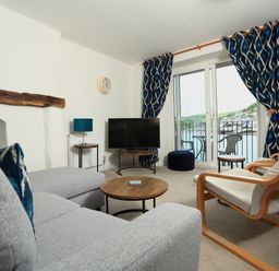 A living room with a gray sofa a wooden armchair a round wooden coffee table a TV on a stand blue patterned curtains and a balcony with chairs overlooking water at Ferry View in Dartmouth