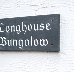 A black rectangular sign with white gothic text reading Longhouse Bungalow on a white textured wall at Longhouse Bungalow in Rock