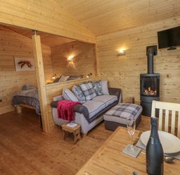 A wooden interior with a sofa bed and ottoman a bed in a corner a fireplace a dining table with a bottle and glass at Foxes Den in Winston near Staindrop