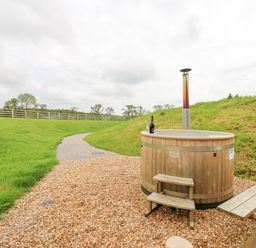 An outdoor wooden hot tub with a chimney and a bottle and glasses on the rim near a wooden picnic table on a gravel path in a grassy field at Foxes Den in Winston near Staindrop
