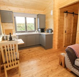 A kitchen with gray cabinets a small dining table with two chairs and a living area with a sofa and wooden sliding door at Otters Holt in Winston near Staindrop