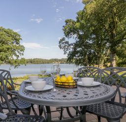 An outdoor metal dining table set with bowls plates wine glasses and a basket of lemons near a lake with trees at Tarn End Cottages 5 Talkin near Brampton Cumbria
