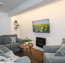 A living room with grey sofas a wall-mounted TV showing sunflowers and a round wooden coffee table at Y Goedwig in Newborough