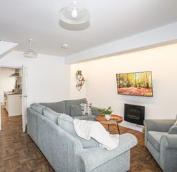 A living room with a grey sectional sofa an armchair a round wooden coffee table a wall-mounted television a potted plant and an open doorway showing a kitchen at Y Goedwig in Newborough