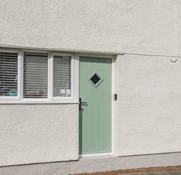 A light green door with a diamond shaped window next to three white framed windows on a white textured wall at Y Goedwig in Newborough