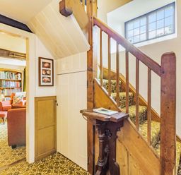 A staircase with patterned carpet and wooden railing next to a hallway with a closed door leading to a living room with bookshelves and armchairs at Saint Pancras Chapel in Washford near Williton