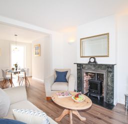 A living room with a wooden coffee table fruit bowl and magazines a beige armchair and sofa a fireplace with a clock and a dining table with chairs in the background at The Shambles in Fortuneswell
