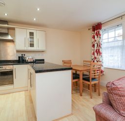 A kitchen with a black countertop island a wooden dining table with four chairs and a window with floral curtains at 1 Tregonwell Court in Minehead