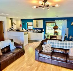 An open living room with two brown leather sofas a wooden dining table with chairs and a kitchen with white cabinets and blue walls at Yr Encil in Llanddaniel near Gaerwen