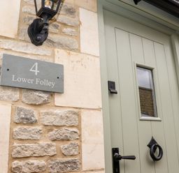 A green wooden front door with a small window and black handle next to a stone wall with a sign reading 4 Lower Folley and a black outdoor light fixture at 4 Lower Folley in Chipping Campden