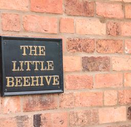 A black sign with gold lettering reading The Little Beehive mounted on a red brick wall at The Little Beehive in Abberley